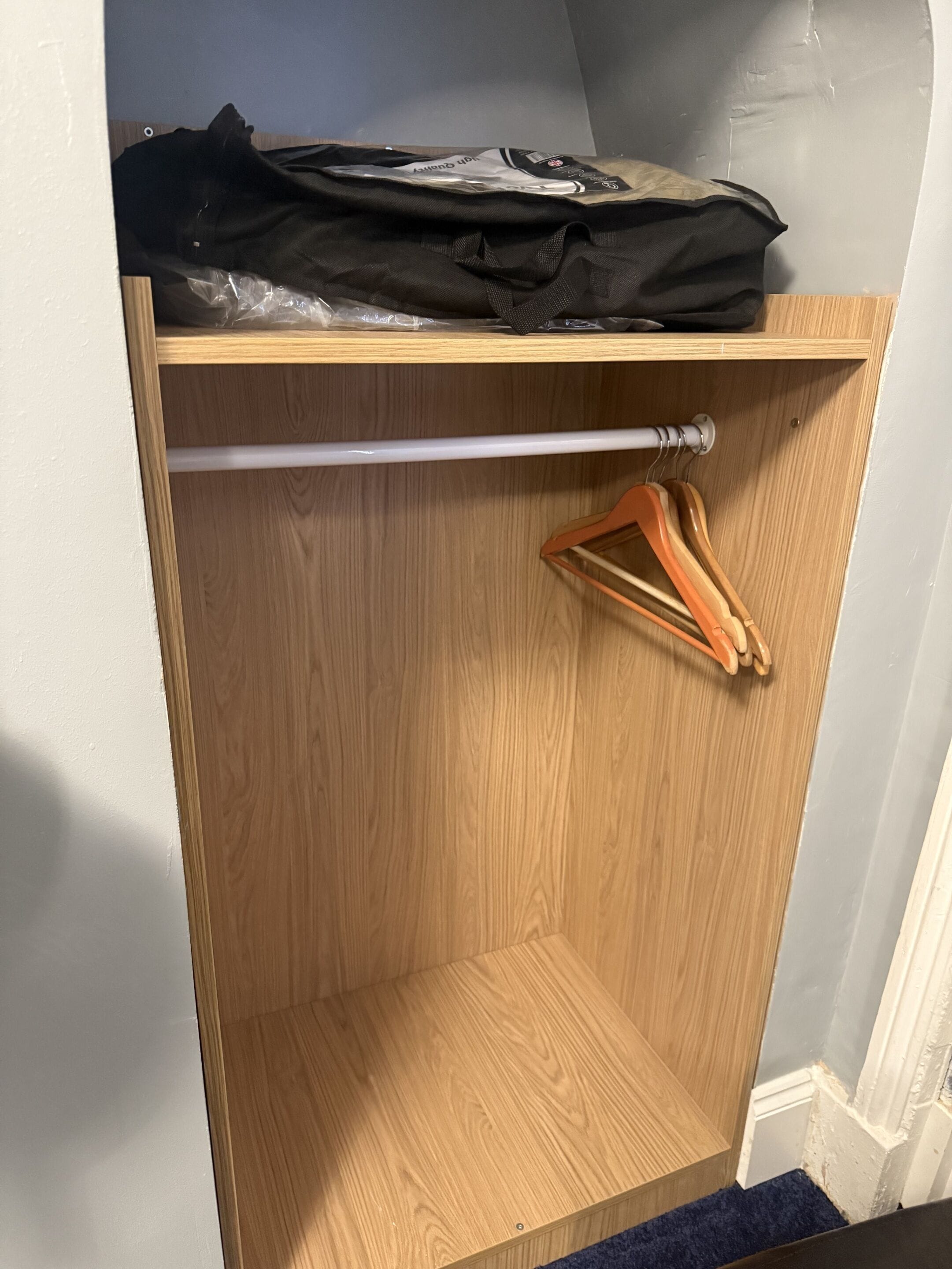 A wooden closet with a hanging rod and a plastic bag on top.
