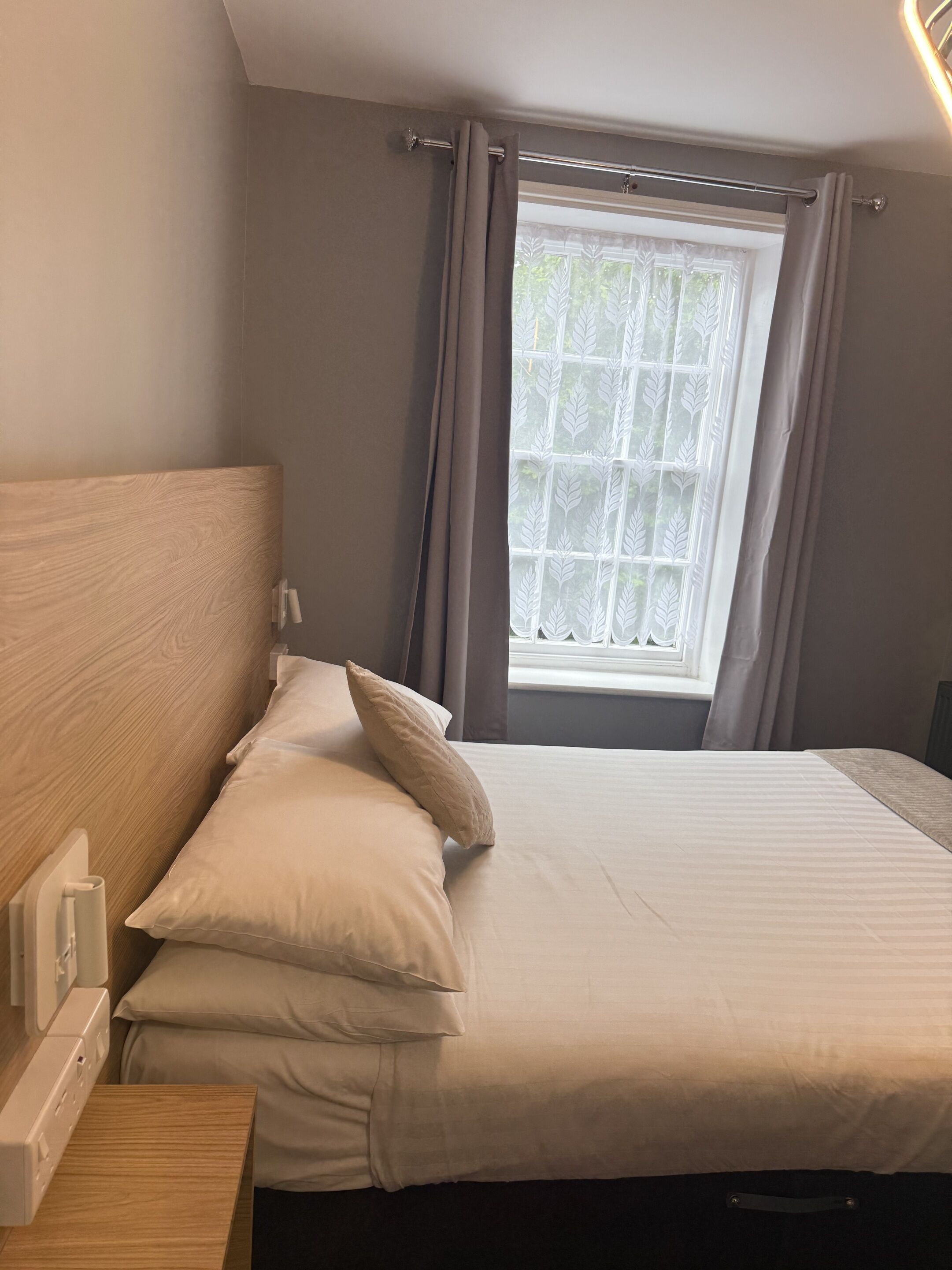 Een slaapkamer met een houten hoofdeinde, een bed met wit beddengoed, een nachtkastje en een raam met gordijnen.
