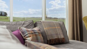 A view from a window with a grey sofa and a plaid blanket, a purple pillow, and a yellow chair. - Elderburn Luxury Lodges (St. Andrews)