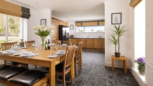 A modern kitchen with wooden cabinets, a dining area with a wooden table set for six, and a potted plant on a small table. - Elderburn Luxury Lodges (St. Andrews)