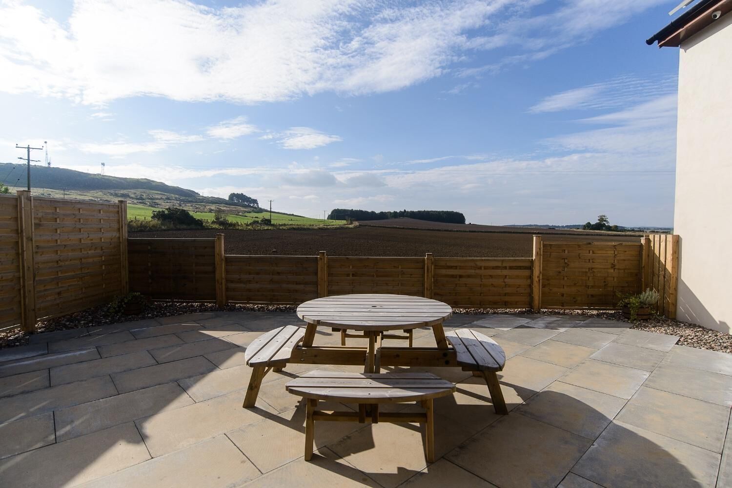 A patio with a wooden table and benches, surrounded by a stone floor and a wooden fence, overlooking a scenic landscape.