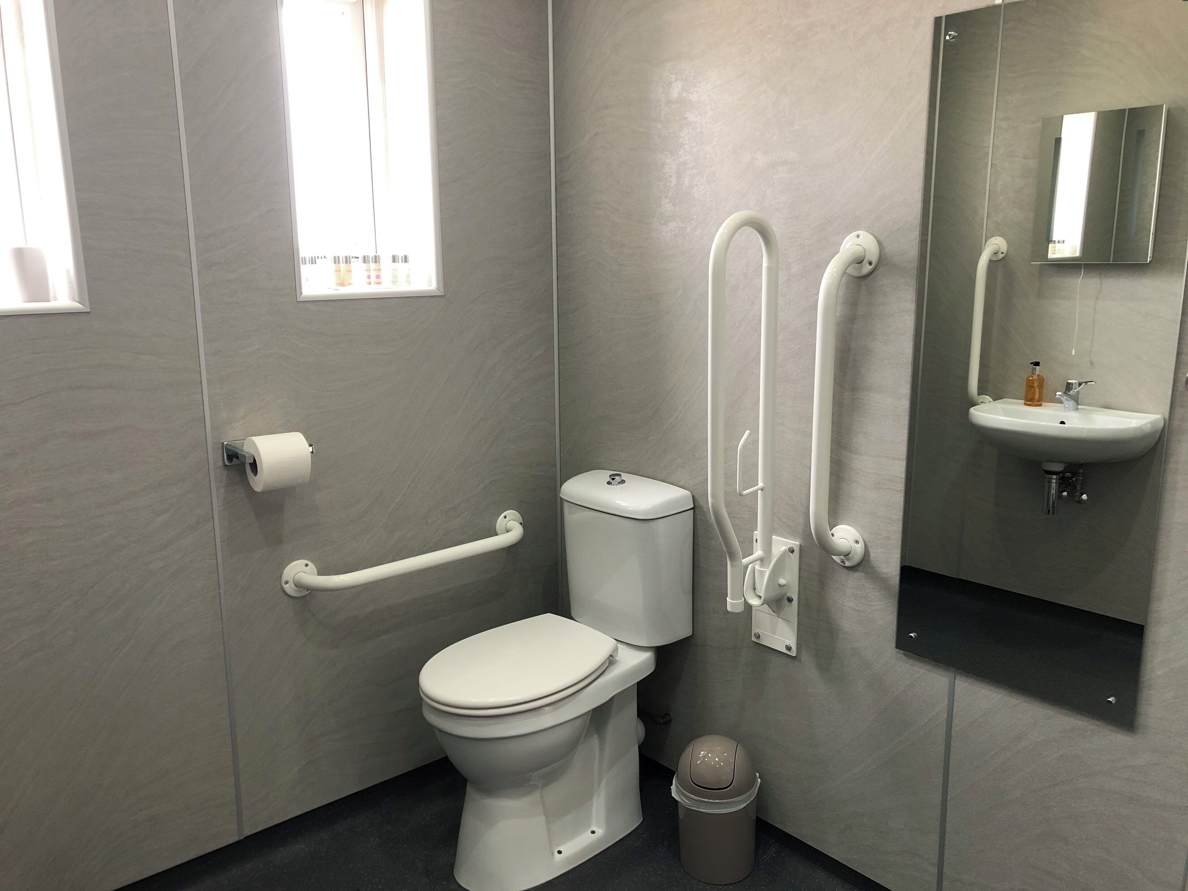 A modern bathroom with a toilet, handrails, a grab bar, a bidet, a sink, and a trash can.