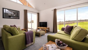 A spacious living room with two green sofas, a wooden coffee table, and a television on a stand. Large windows offer a view of the outdoors. - Elderburn Luxury Lodges (St. Andrews)