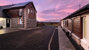 A residential street with houses featuring stone and wooden exteriors, a gravel driveway, and a sunset sky. - Elderburn Luxury Lodges (St. Andrews)