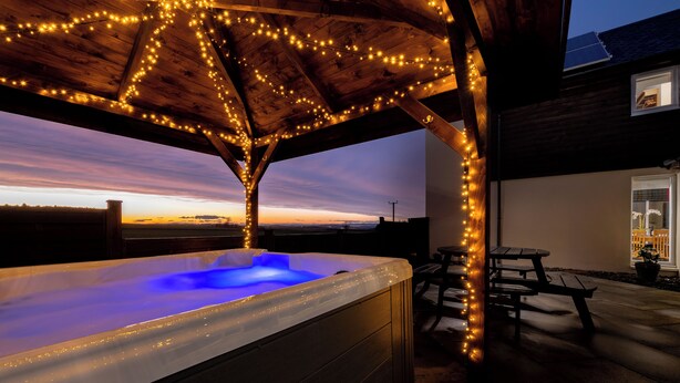 A hot tub under a wooden pergola with string lights, overlooking a sunset view. - Elderburn Luxury Lodges (St. Andrews)