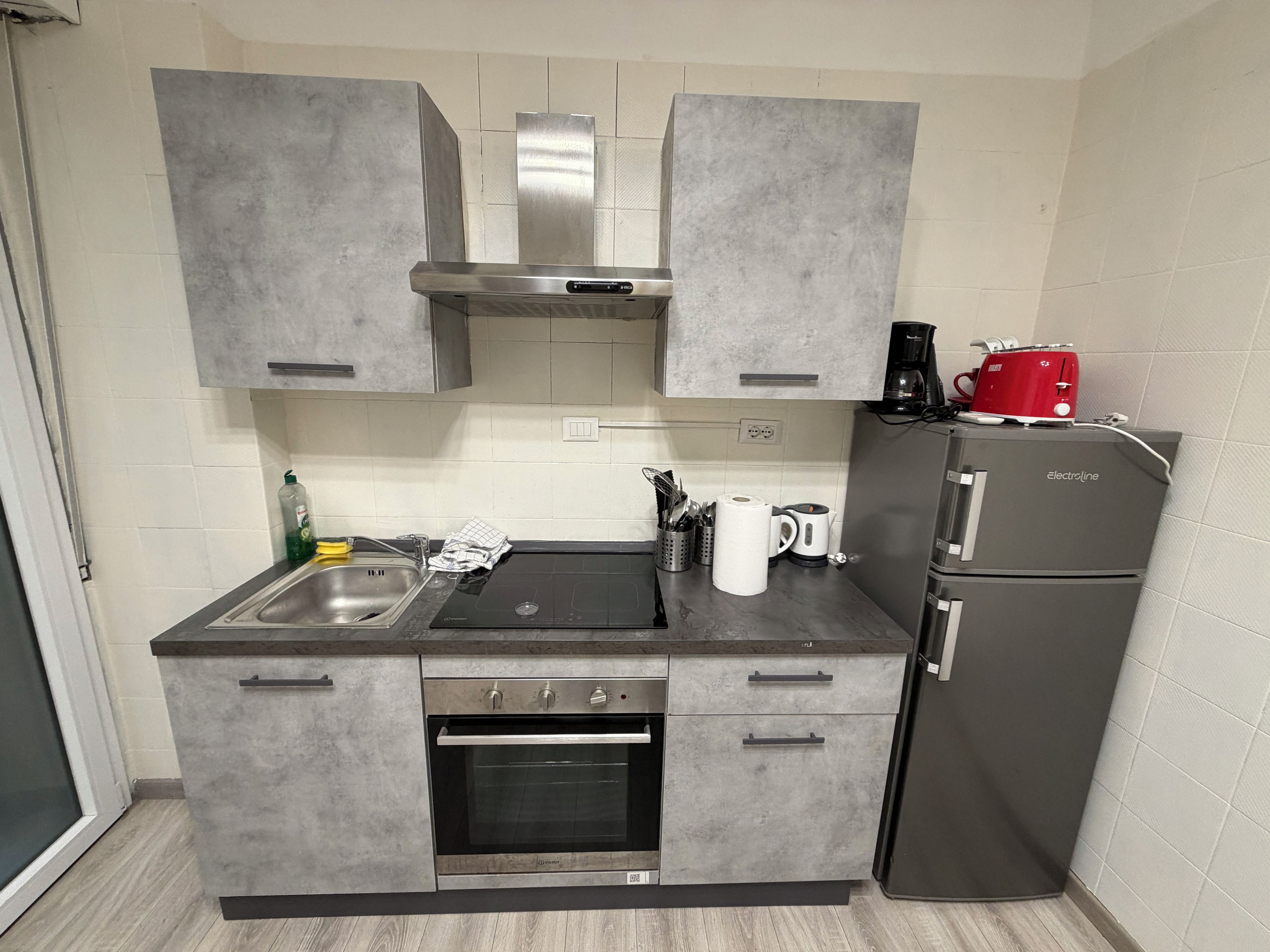 Una cocina compacta con gabinetes grises, horno empotrado, lavamanos y refrigerador.
