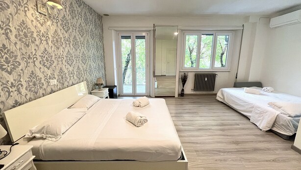 A hotel room with a large bed, a smaller bed, a desk, and a chair. There is a window with a view of trees and a door leading to a balcony. - A Quattro Passi Da (Rome)