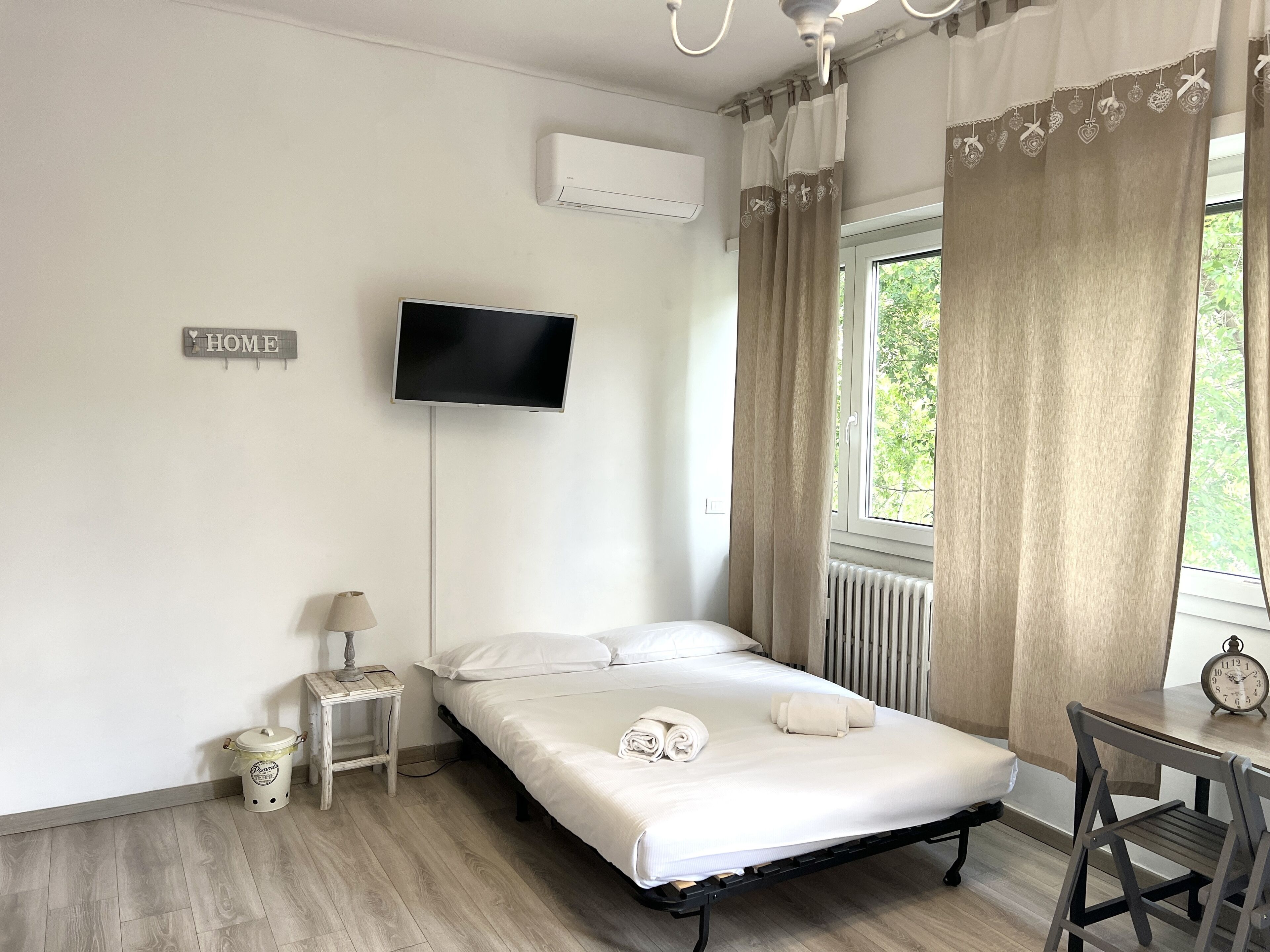 Ein Schlafzimmer mit einem Bett, einem Fernseher, einem Heizkörper, einem Fenster mit Vorhängen, einem kleinen Tisch und einem Stuhl.