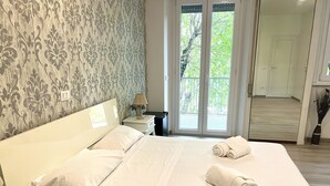 A hotel room with a large bed, a nightstand, a lamp, and a view of a balcony with greenery. - A Quattro Passi Da (Rome)