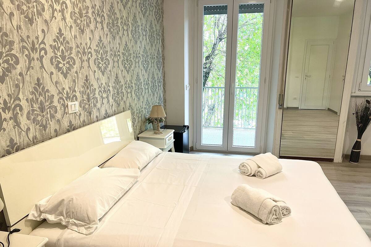 A hotel room with a large bed, a nightstand, a lamp, and a view of a balcony with greenery.