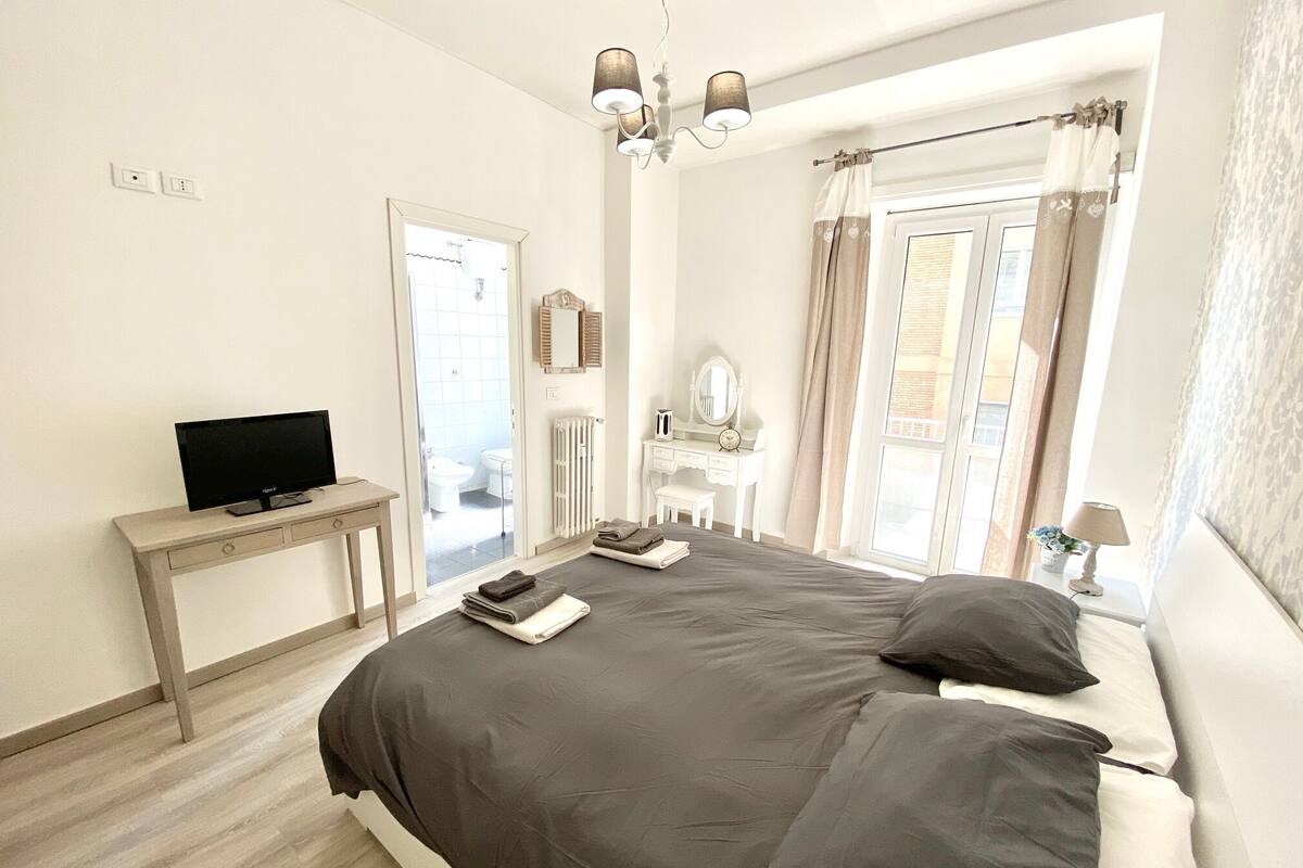 A bedroom with a bed, a TV on a side table, a dresser, a mirror, and a window with curtains.