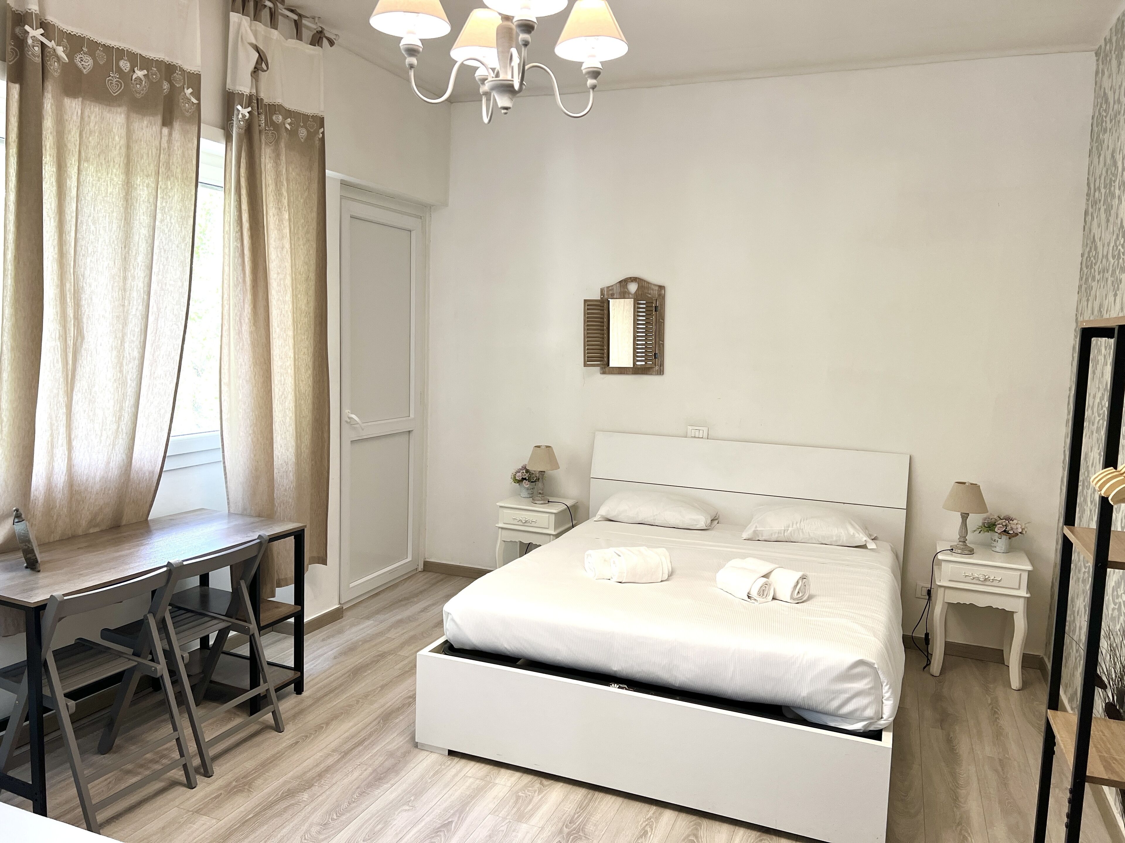 A bedroom with a bed, a desk, and a chair. There is a chandelier above the bed and a mirror on the wall.