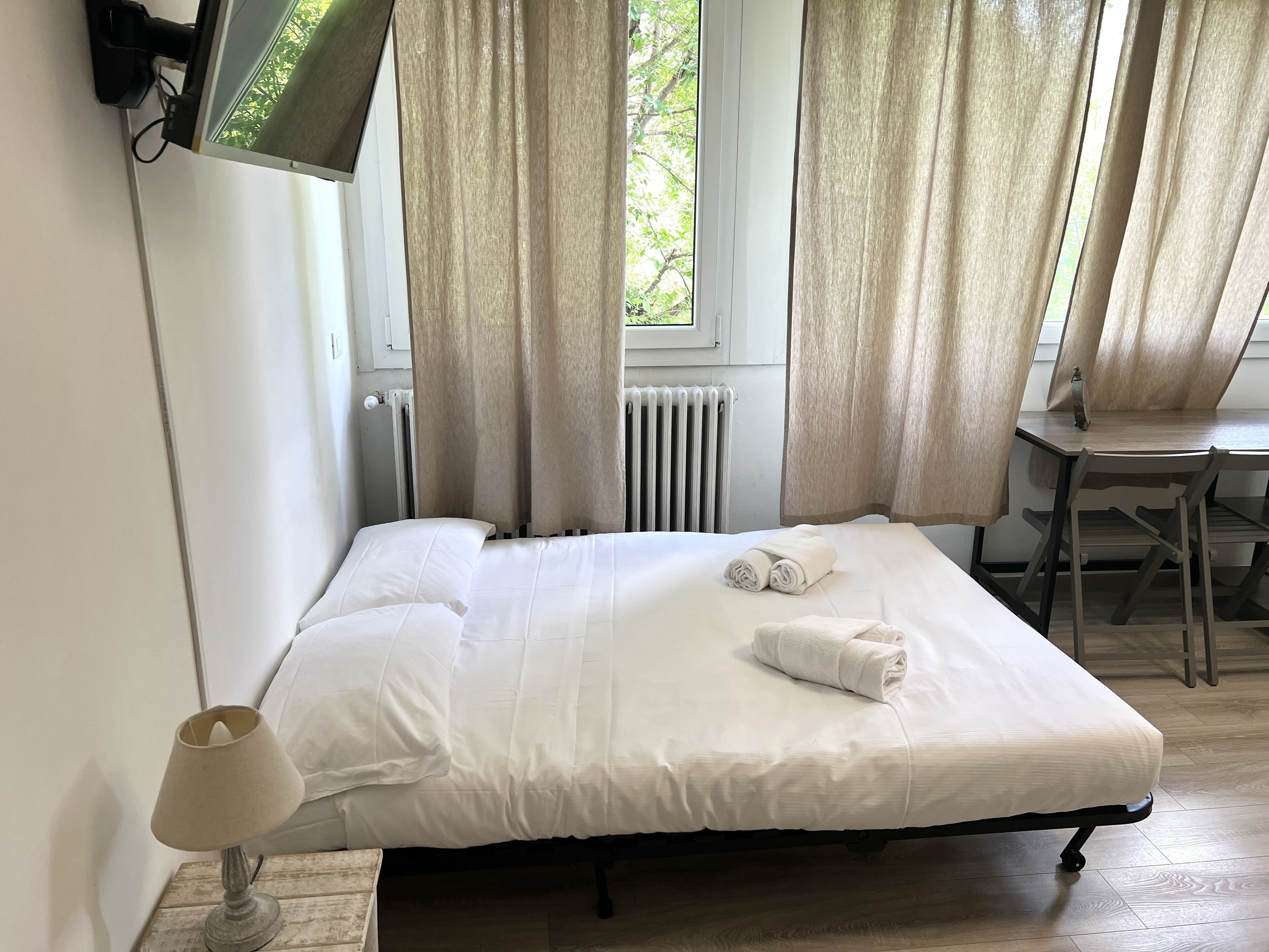 A neatly made bed with white linens and a rolled towel, a wooden desk with a chair, a television mounted on the wall, and a window with curtains.