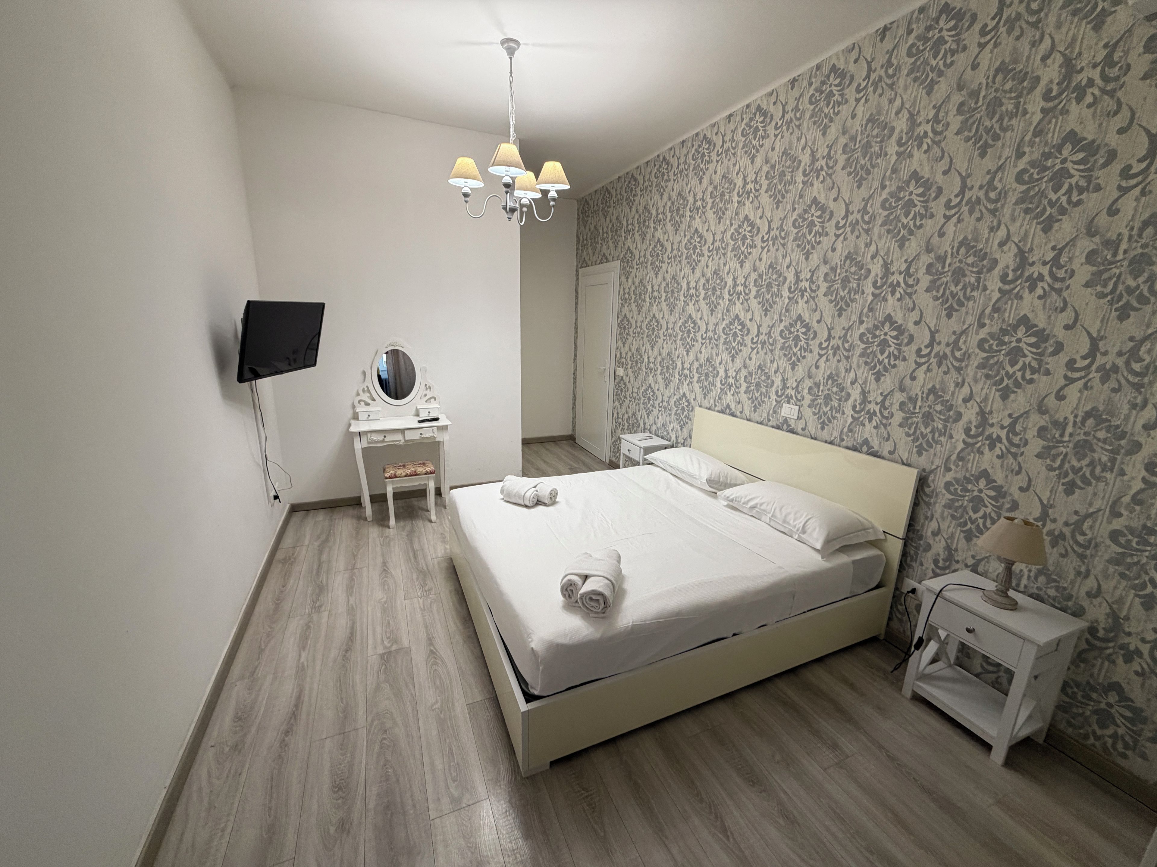 Quarto com cama, penteadeira com espelho, mesa de cabeceira e televisão fixada na parede.