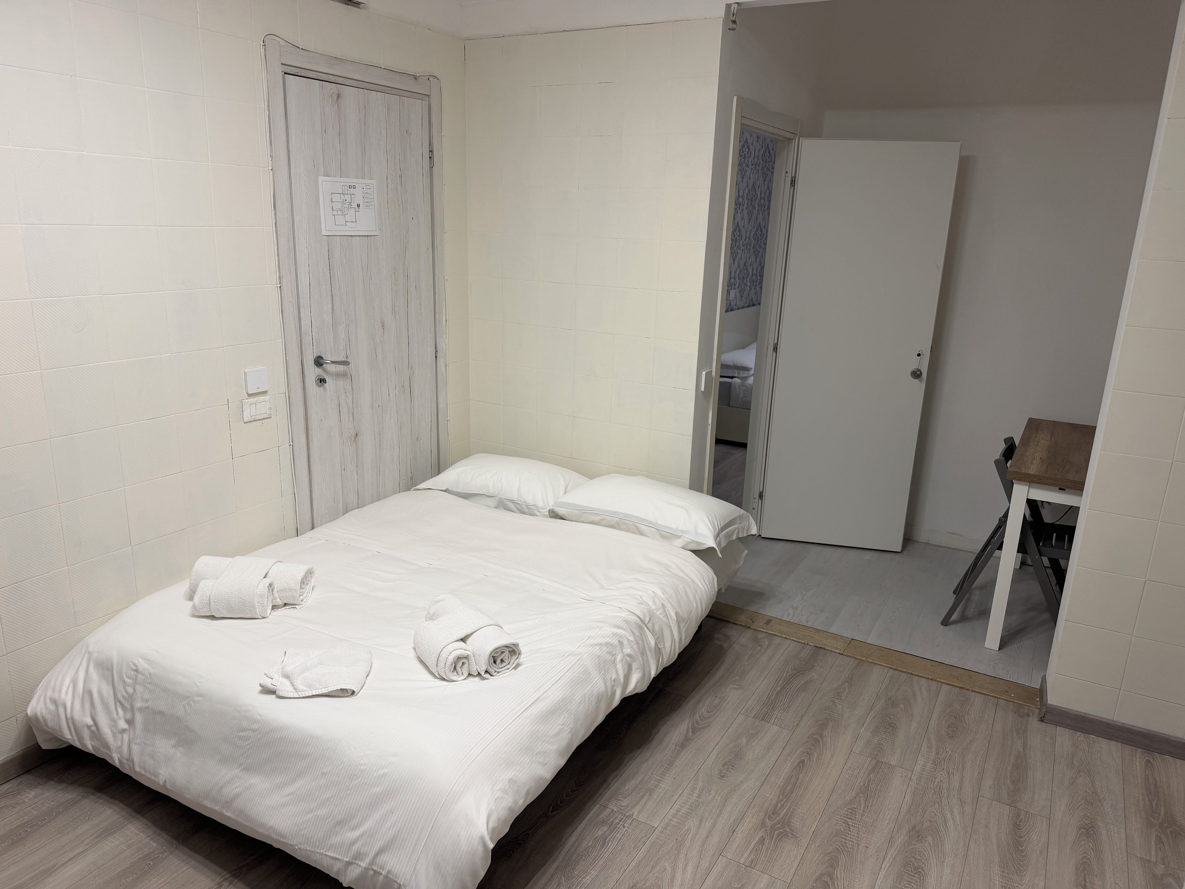 A single-bed room with a wooden floor, a white door, and a small table.