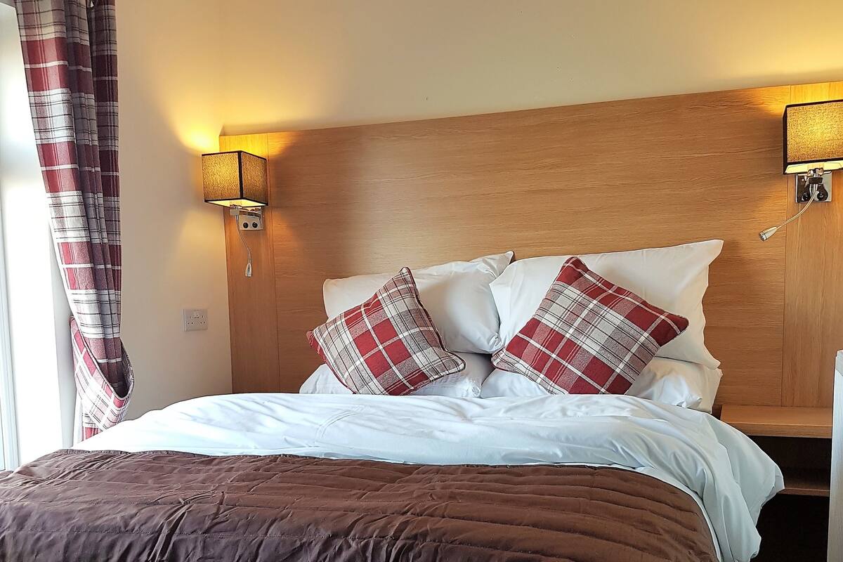 A neatly made bed with a brown comforter and plaid pillows, flanked by two wall-mounted lamps, in a room with wooden paneling.