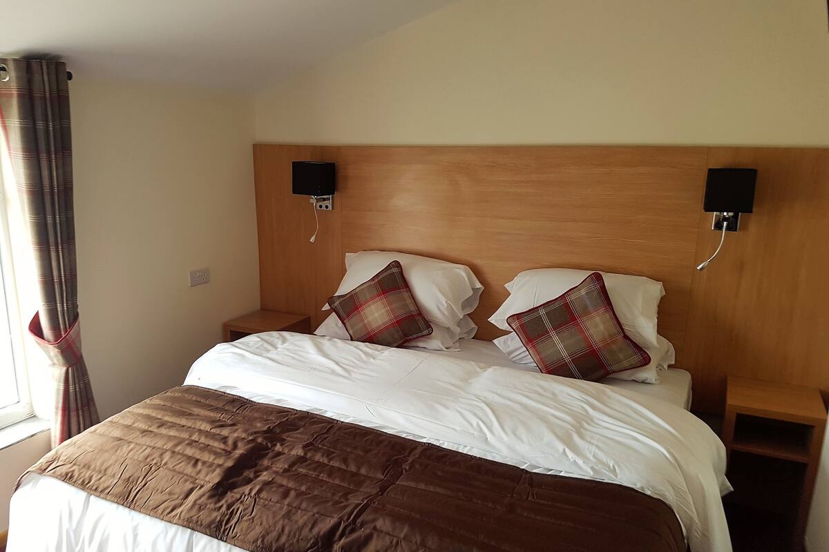 A neatly made bed with a brown quilt, white pillows, and plaid throw pillows. There are two bedside tables with lamps, and the room has a wooden headboard.
