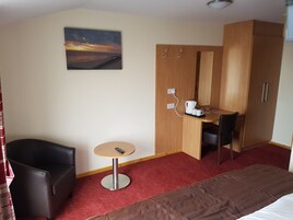 A hotel room with a bed, a small round table with a remote control, a desk with a coffee maker, a chair, a wardrobe, and a painting on the wall.