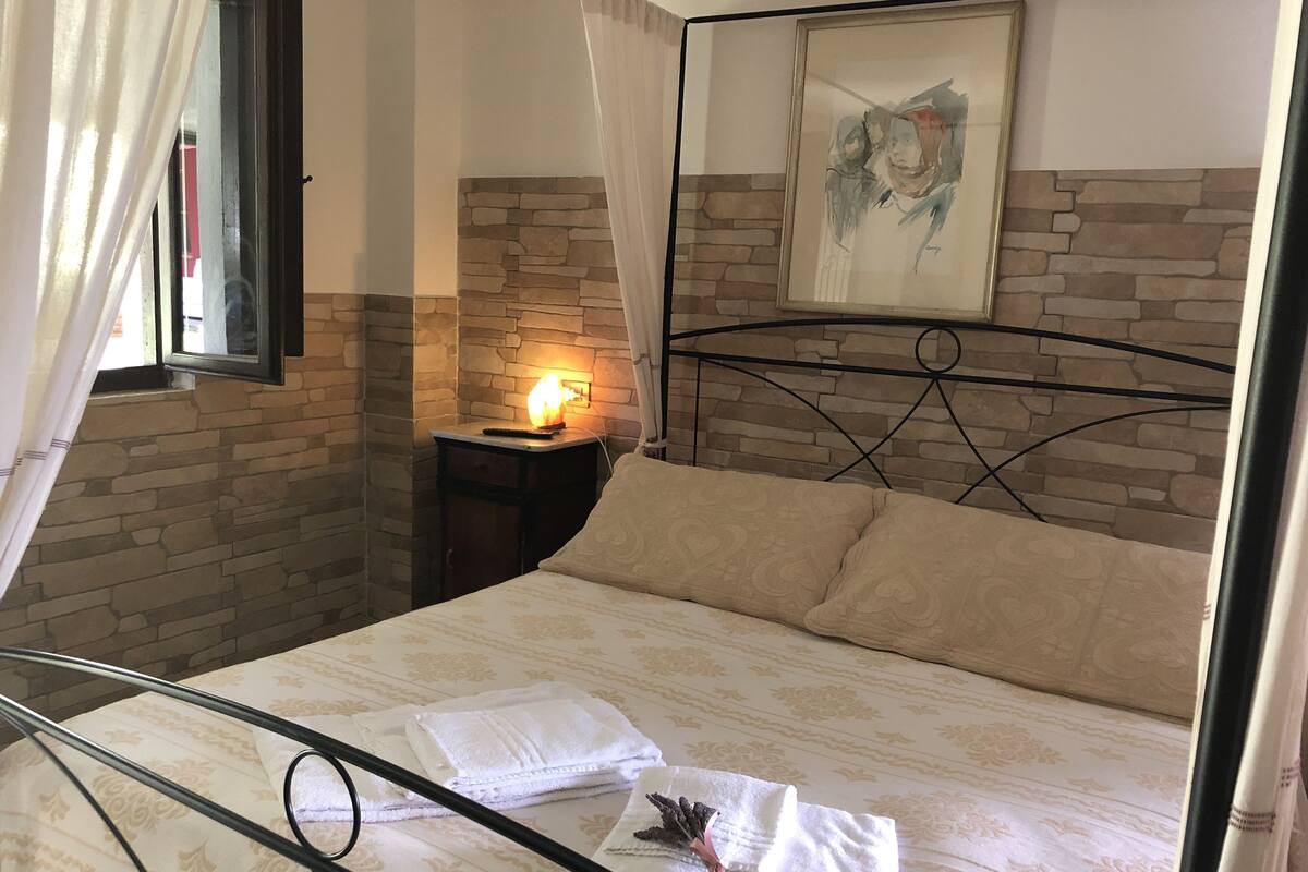 Un dormitorio con cama, mesita de noche, lámpara, ventana con cortinas, pared de piedra y un cuadro en la pared.