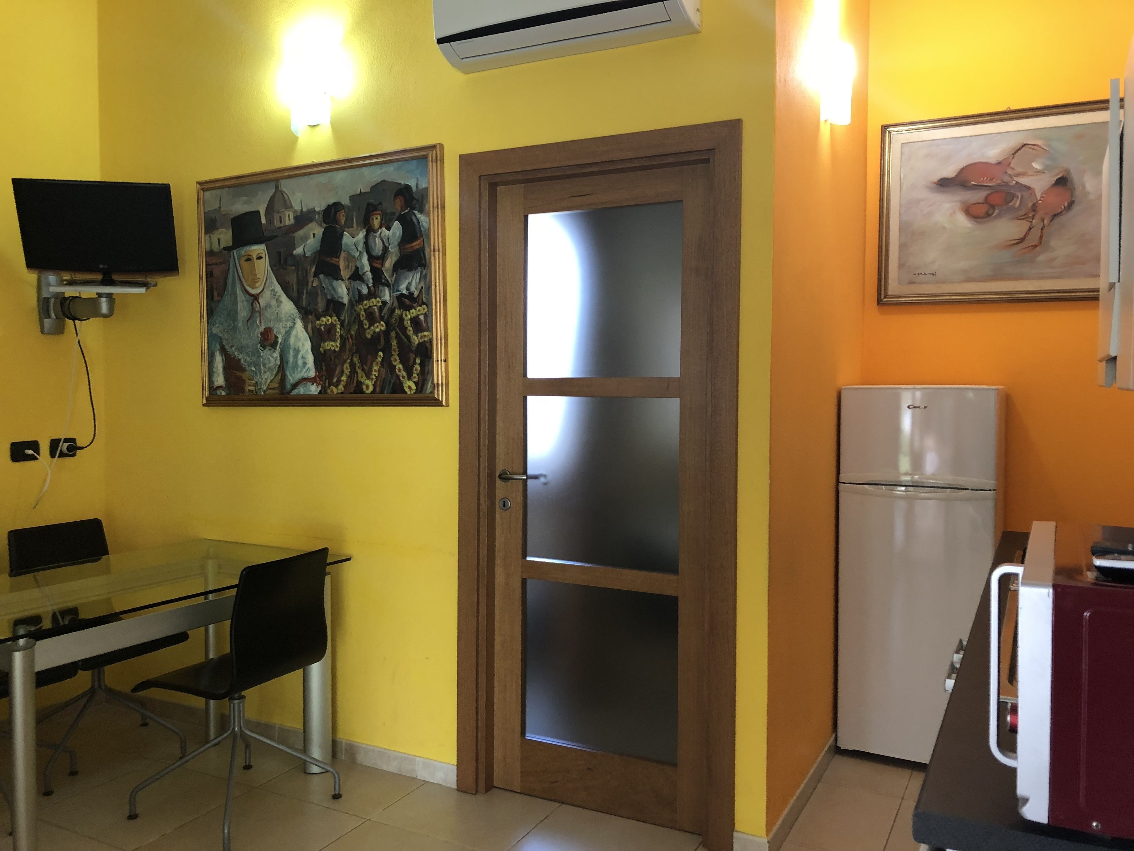 Una cocina funcional con una mesa de vidrio, sillas, un refrigerador y dos cuadros en las paredes.