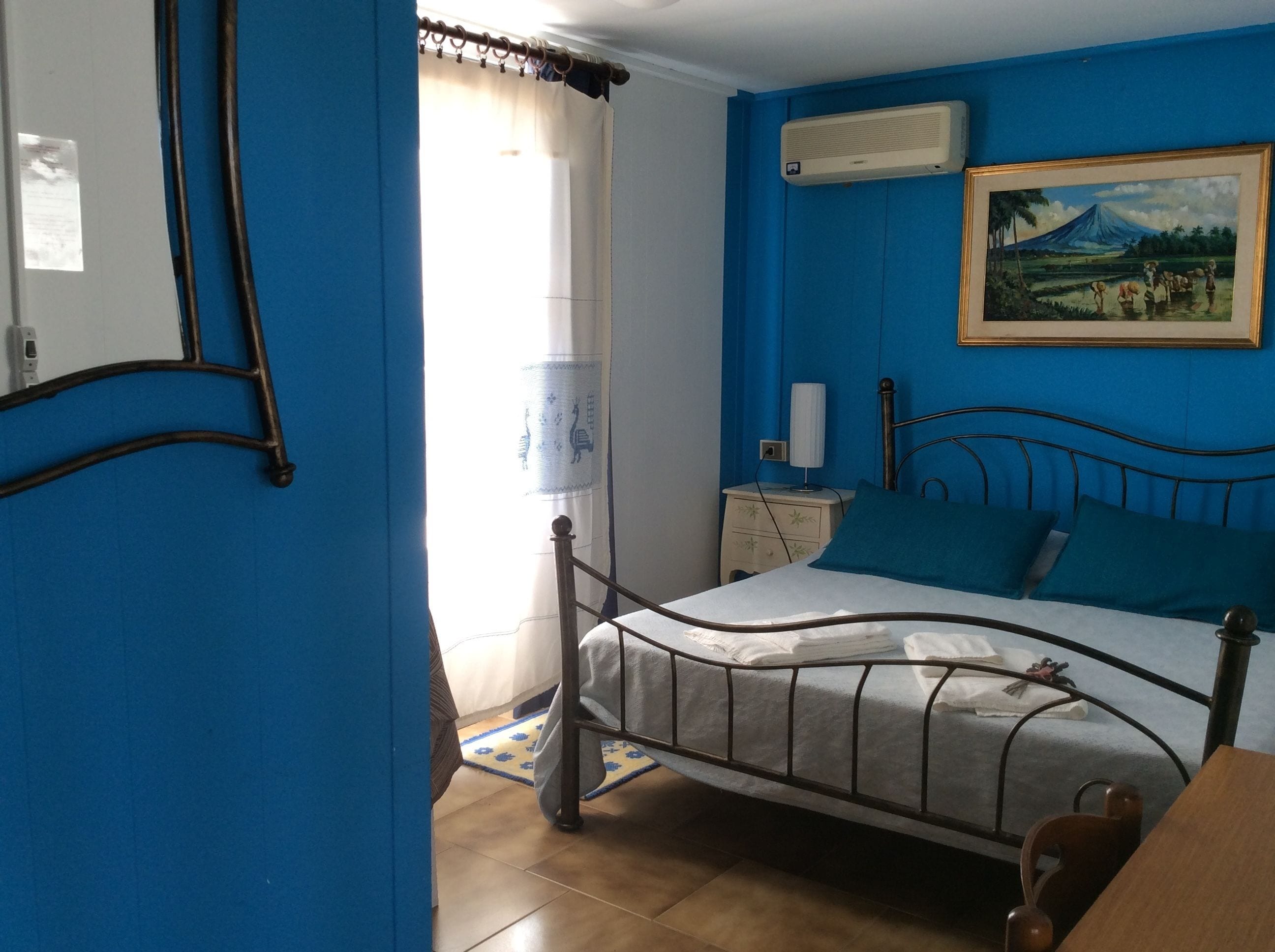 Un dormitorio con cama, mesita de noche, lámpara, cuadro y ventana con cortinas.