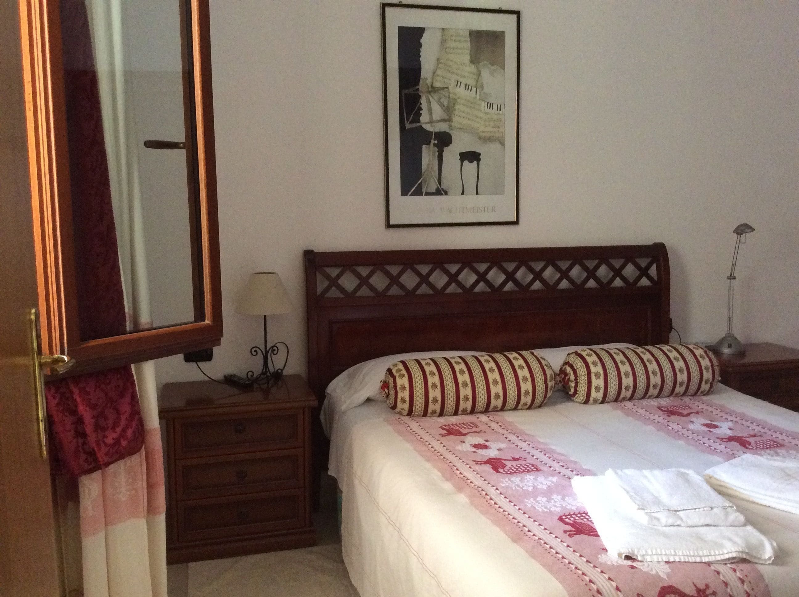 Een slaapkamer met een houten hoofdbord, een bed met beddengoed met een patroon, een nachtkastje met een lamp, een spiegel en een lijst met een afbeelding aan de muur.