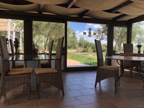 Uma sala de jantar com cadeiras de vime e uma mesa de vidro, janelas amplas com vista para um jardim e árvores.