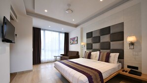 Ein modernes Hotelzimmer mit einem großen Bett, einem Flachbildfernseher, einer Sitzecke mit Sessel und Tisch sowie einem großen Fenster mit Vorhängen.