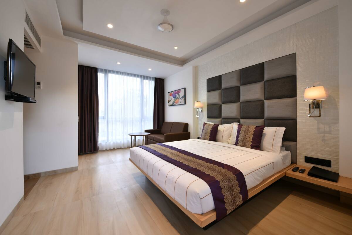 Una habitación de hotel moderna con una cama grande, un televisor de pantalla plana, una zona de estar con una silla y mesa, y un ventanal con cortinas.