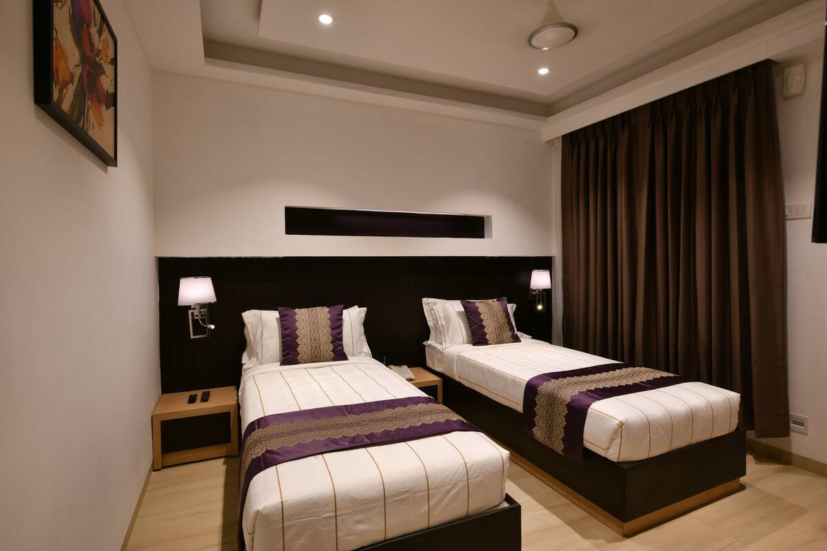 Habitación de hotel con dos camas, una mesita de noche, una lámpara de pared y un ventanal con cortinas.