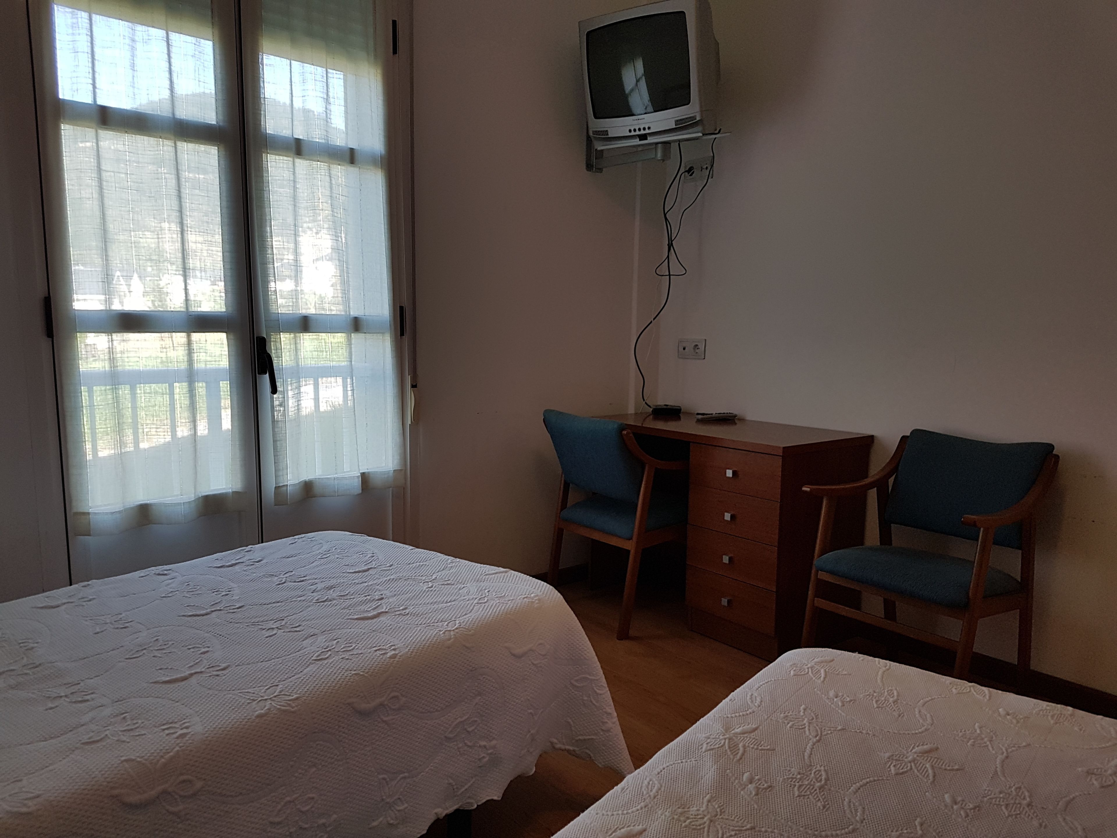 Una habitación con dos camas, un escritorio con silla, un televisor montado en la pared y una ventana con cortinas.