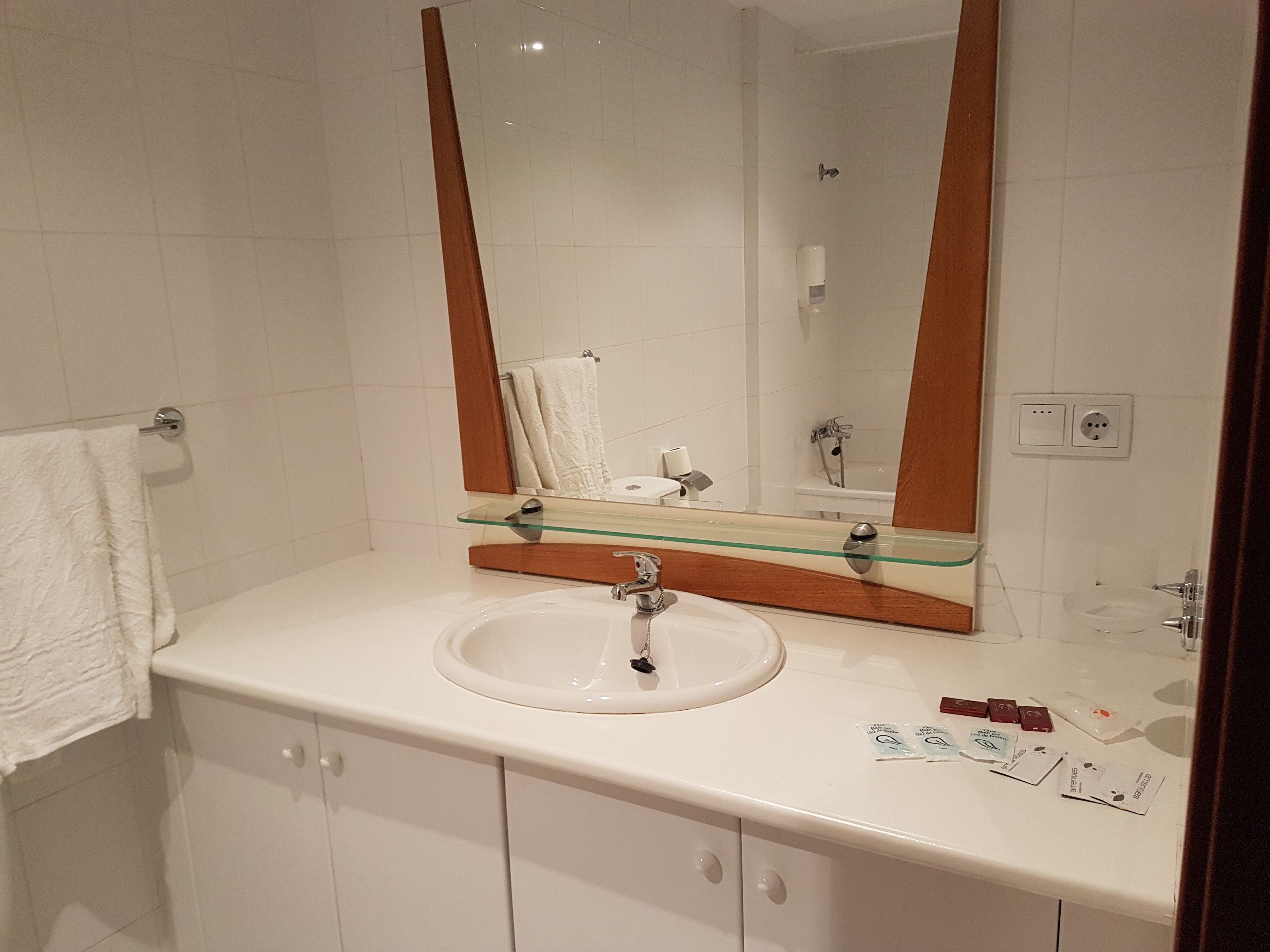 Un baño con un lavamanos blanco, un estante de vidrio arriba y un espejo grande.