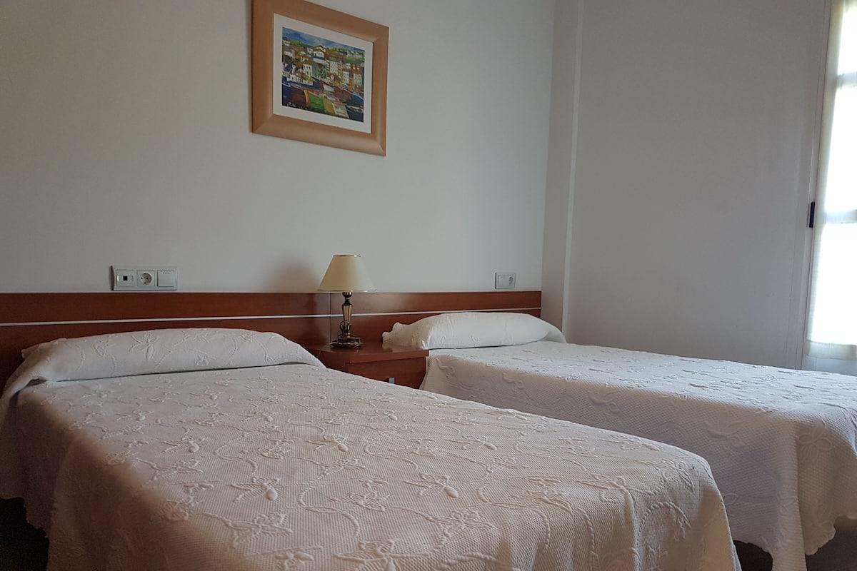 A hotel room with two single beds, a nightstand with a lamp, a painting on the wall, and a window with curtains.