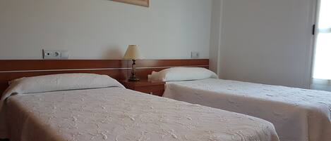 Habitación de hotel con dos camas individuales, una mesita de noche con lámpara, un cuadro en la pared y una ventana con cortinas.