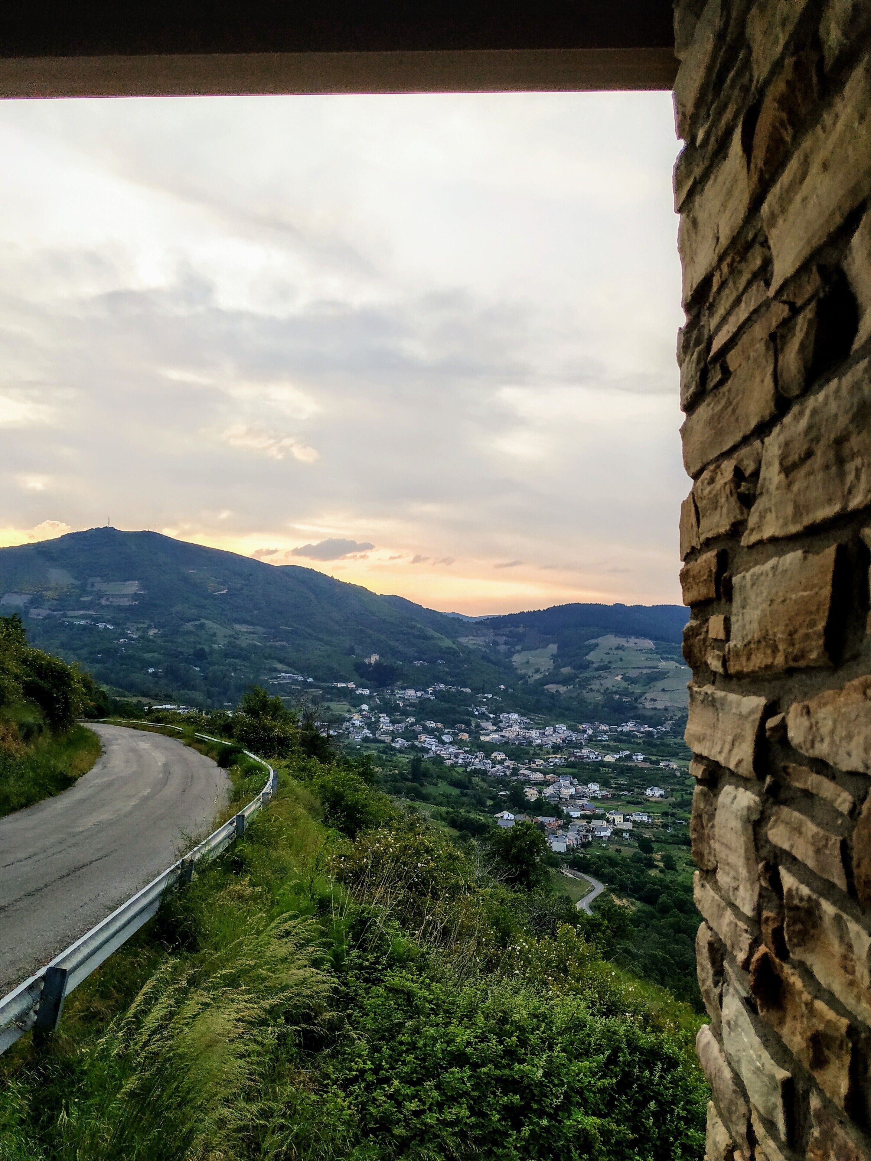 Un camino sinuoso con un muro de piedra a la derecha y vistas de un valle con casas.