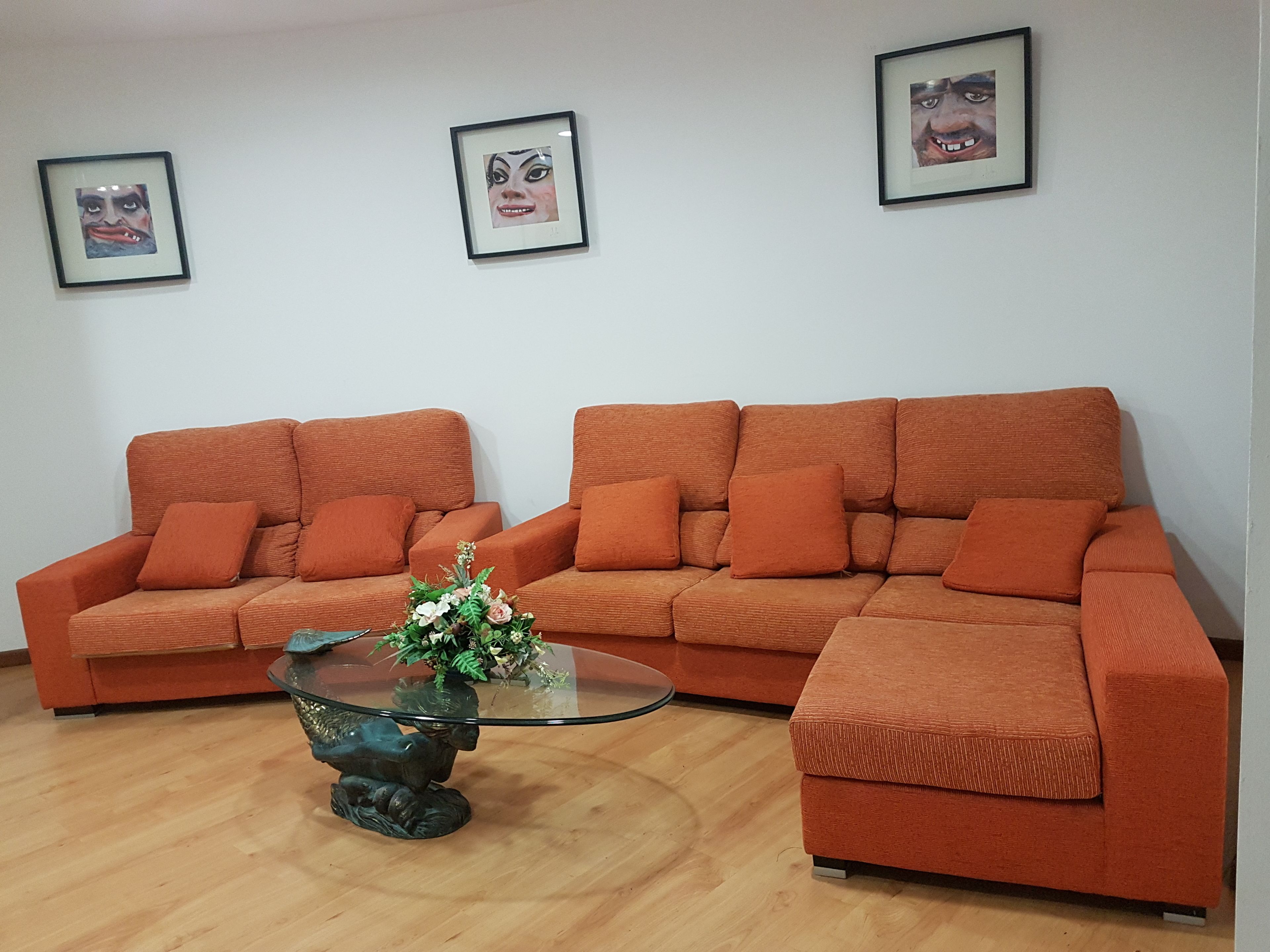 A living room with two orange sofas, a glass coffee table with a floral centerpiece, and three framed pictures on the wall.