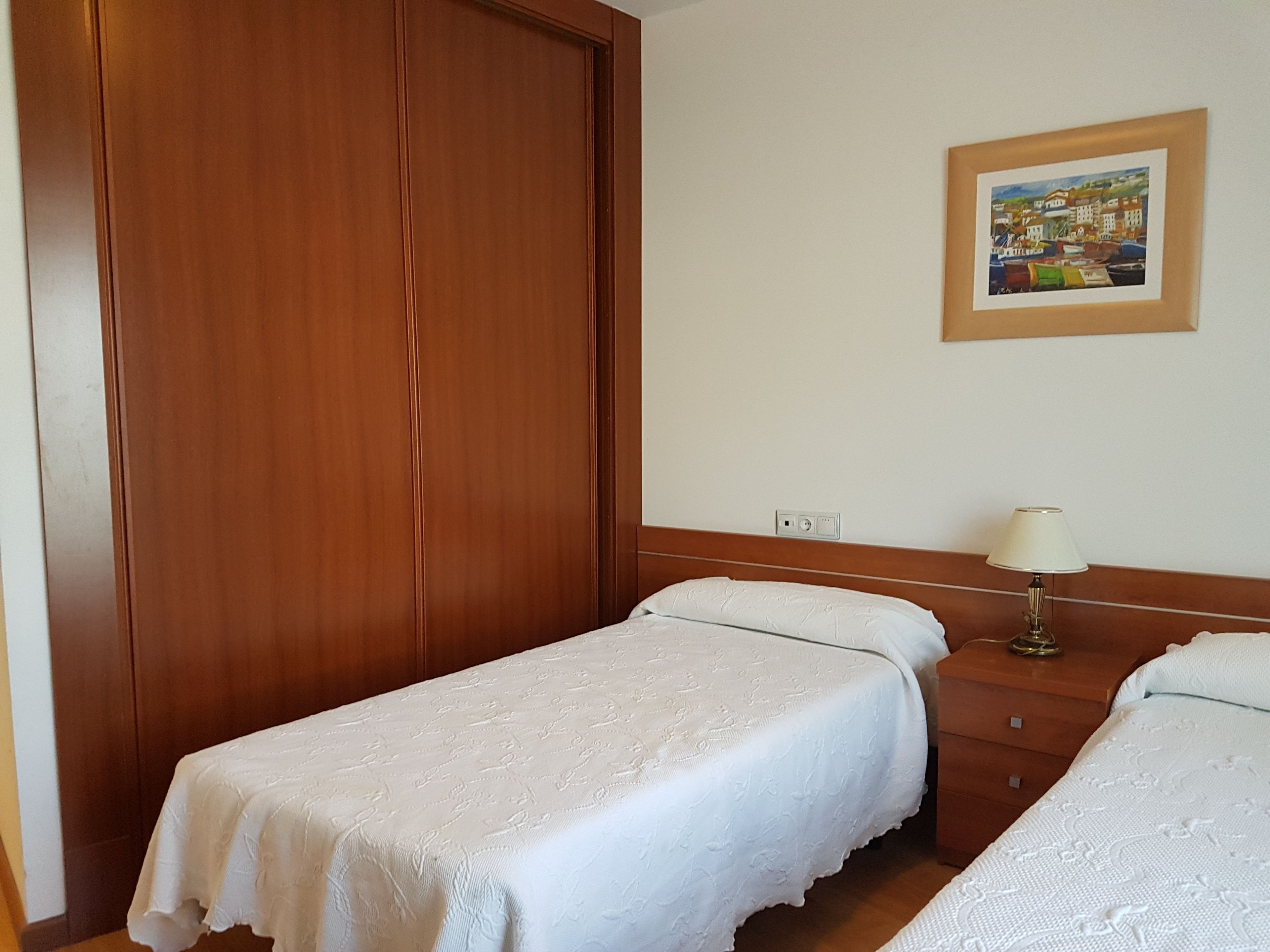 Habitación de hotel con dos camas individuales, un armario de madera, una mesita de noche con lámpara y un cuadro enmarcado en la pared.