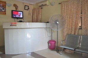 A reception area with a tiled floor, a reception desk, a television mounted on the wall, a standing fan, a pink trash bin, and a bench. - Kings Crown Hotel (Lagos)