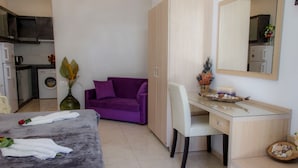 Un dormitorio con una cama, un sofá púrpura, un escritorio con un espejo y una pequeña kitchenette al fondo.