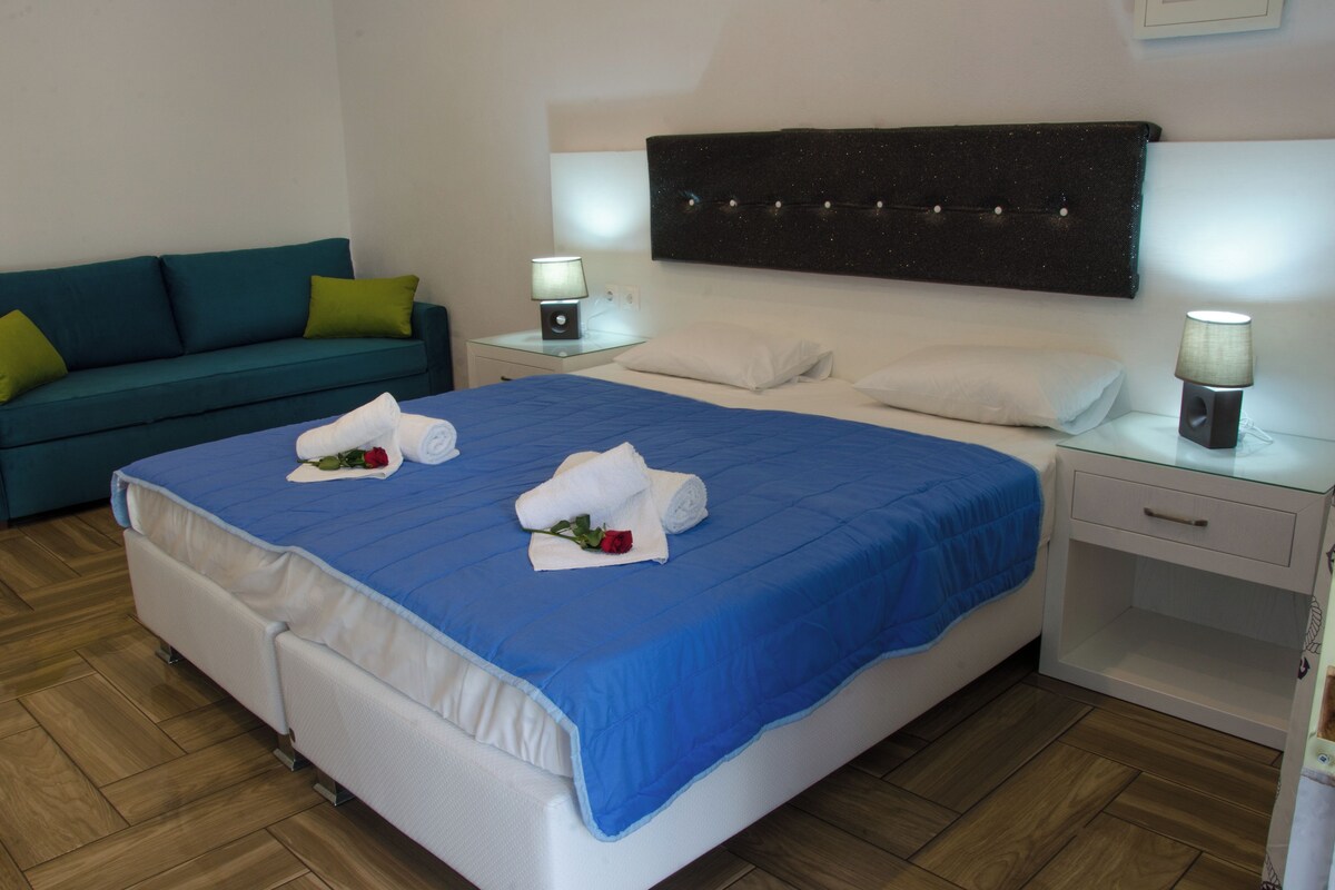 Habitación de hotel con una cama grande, una mesita de noche con lámpara, un sofá con cojines y un cabecero fijado a la pared.