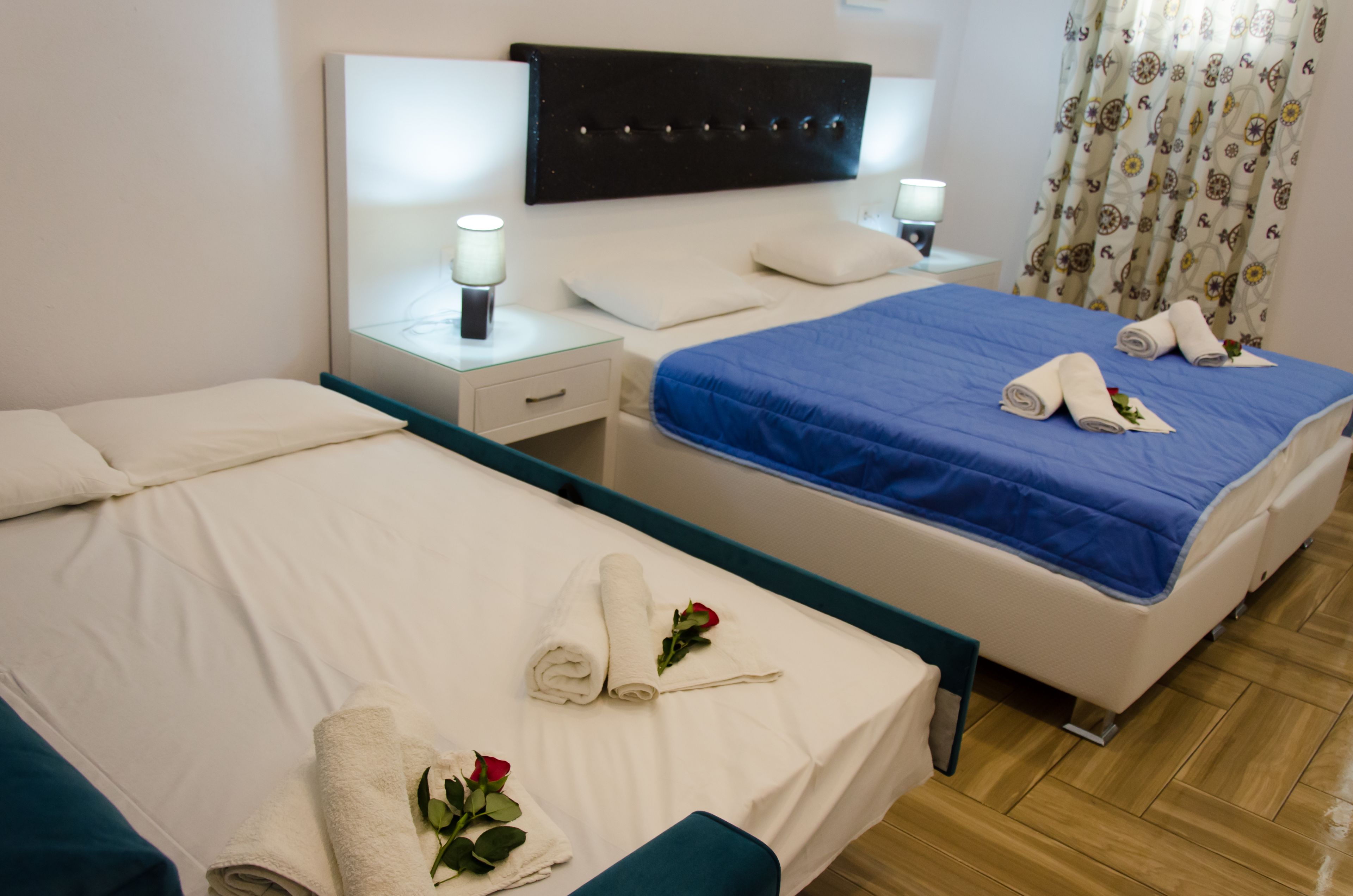 Habitación de hotel con dos camas, cada una con cabecera, ropa blanca y una colcha azul en una de ellas. Hay toallas enrolladas y una rosa sobre las camas.