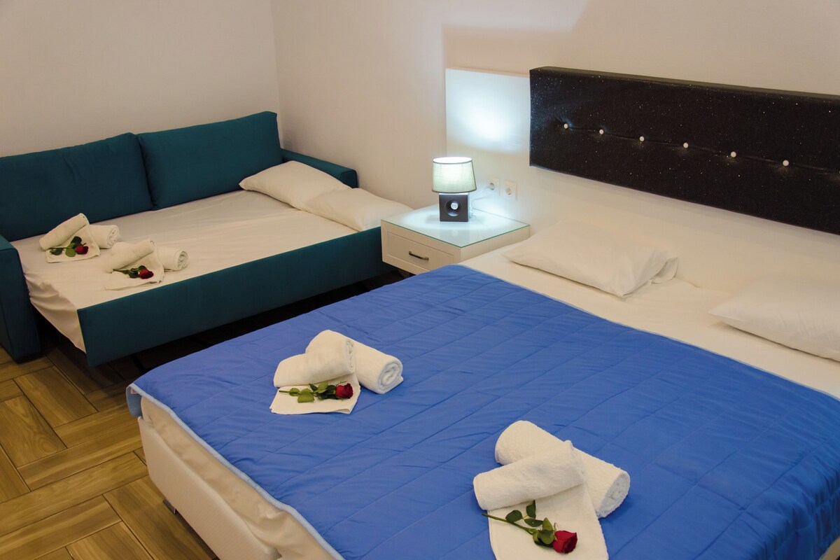 Habitación de hotel con dos camas, una con cubrecama azul y la otra con cubrecama verde azulado. Cada cama tiene una toalla enrollada y una rosa. Entre las camas hay una mesita de noche con una lámpara.