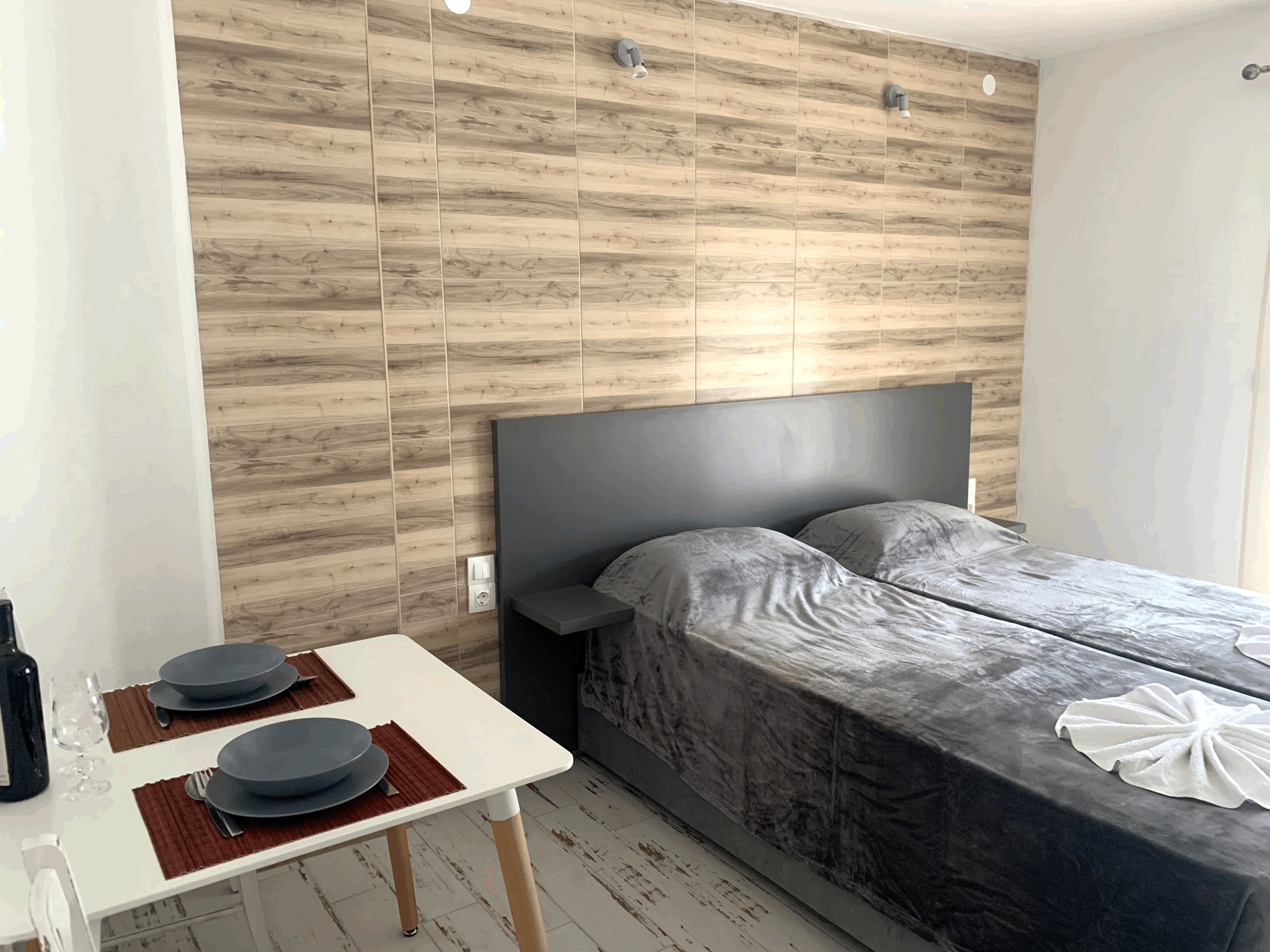 A hotel room with a bed, a small table with plates and cutlery, a wall with wooden panels, and a light fixture.