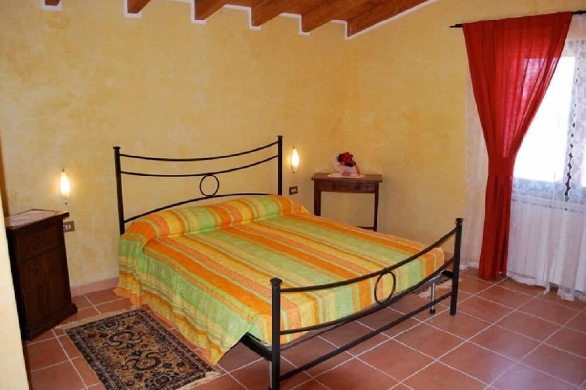 Una cama individual con estructura metálica, un tapete estampado, una mesita de noche pequeña y una ventana con cortinas rojas.