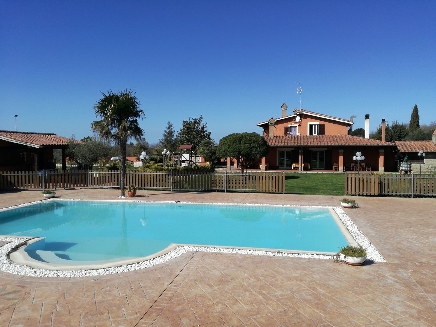 Una piscina con bordo piastrellato, circondata da un recinto in legno e un edificio con tetto in tegole.