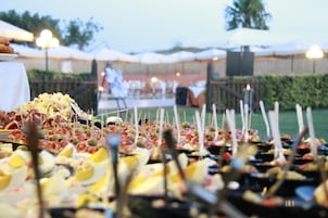 An outdoor buffet with various food items on skewers, set up under tents with a stage in the background.