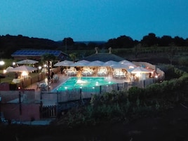 A poolside area with multiple tables and umbrellas, surrounded by a stone wall and vegetation.