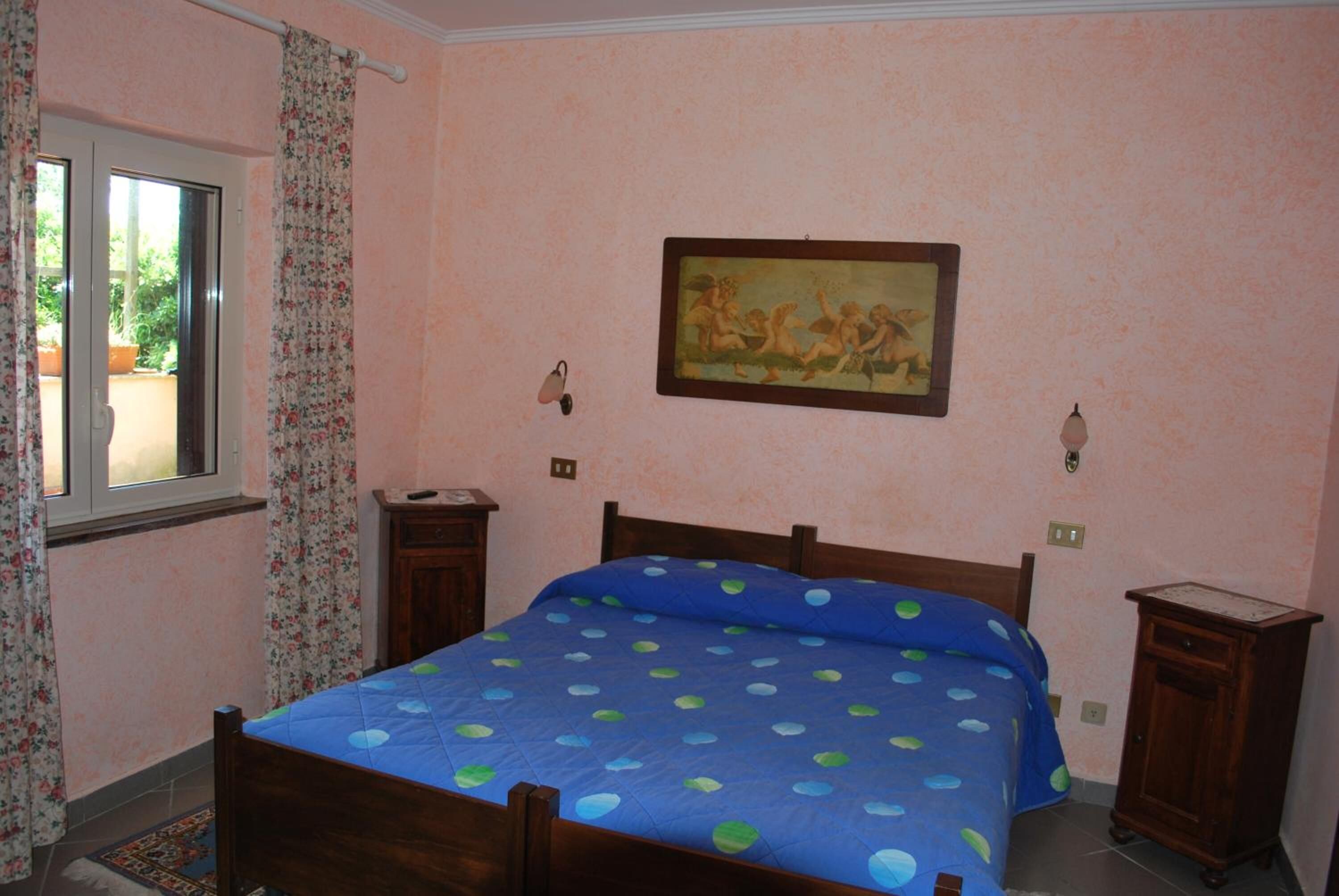 Een slaapkamer met een bed, een raam met gordijnen, een schilderij aan de muur en een klein kastje.