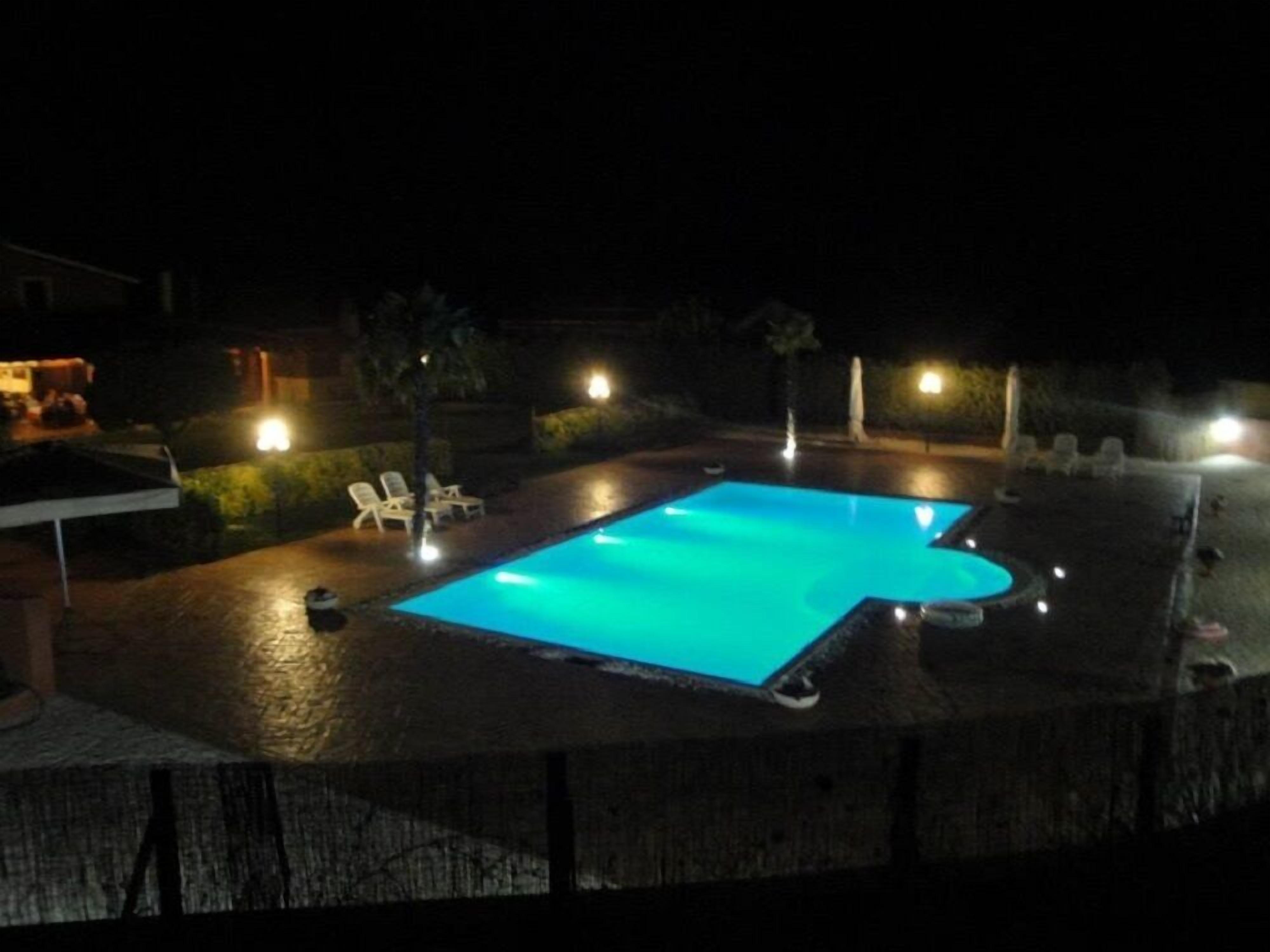 A swimming pool at night with surrounding lounge chairs and a covered area.