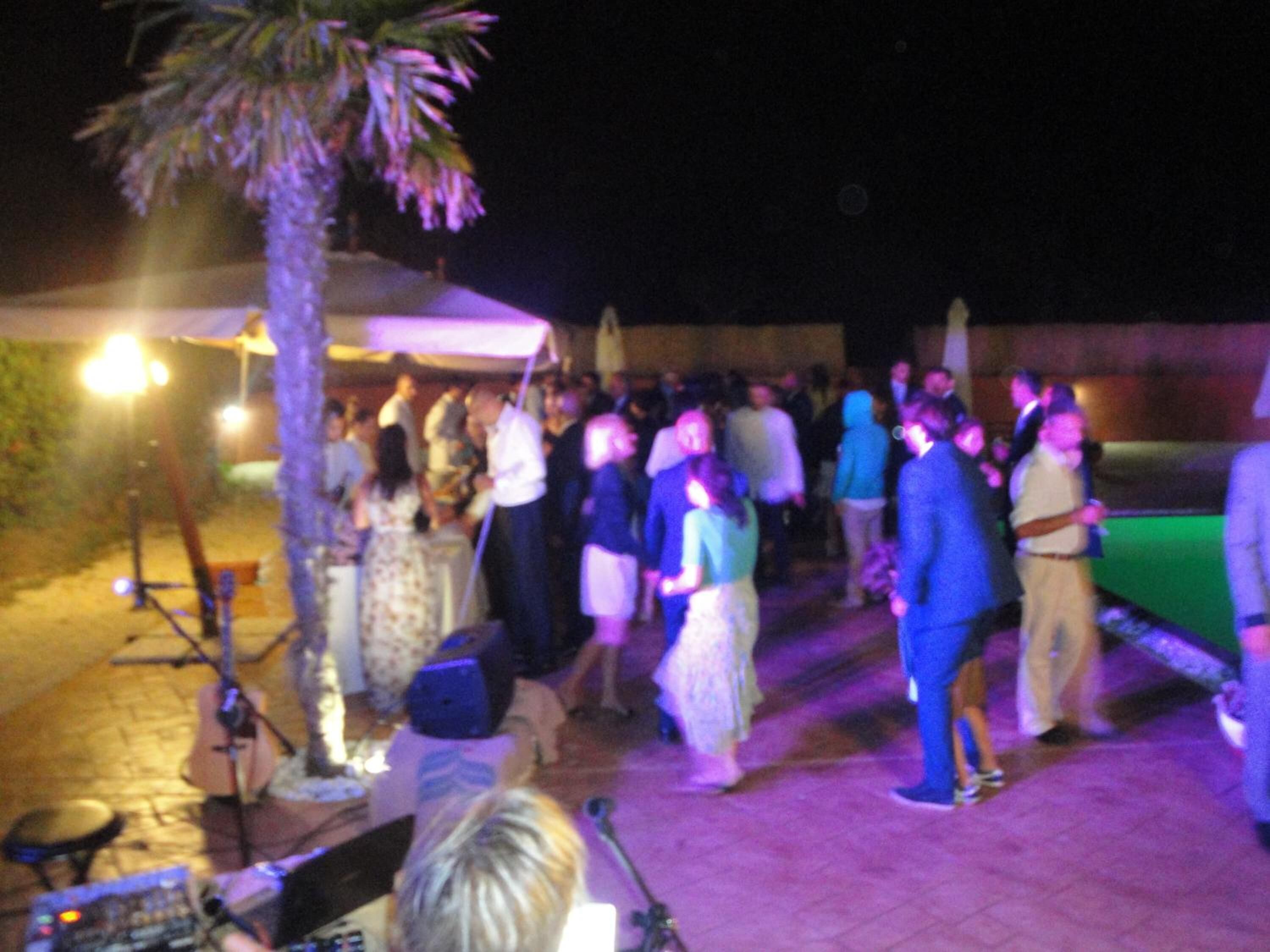 Een druk evenement buiten met mensen die samenkomen, een podium met muziekinstrumenten en een palmboom.